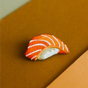 以下が含まれることがあります： サーモンの寿司の刺繍パッチのクローズアップ。パッチはオレンジと白で、サーモンの握りの詳細なデザインが施されています。
