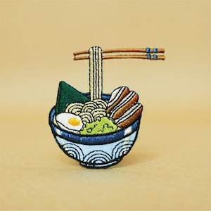 以下が含まれることがあります： ラーメンの刺繍パッチ。麺、卵、海苔、肉が入ったラーメンの入った青い波模様の丼。丼から箸が2本出ている
