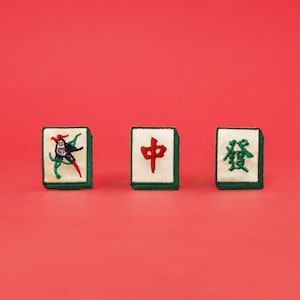 May include: Three green and white mahjong tiles with embroidered designs. The first tile features a bird, the second tile has the Chinese character for "middle", and the third tile has the Chinese character for "bamboo".