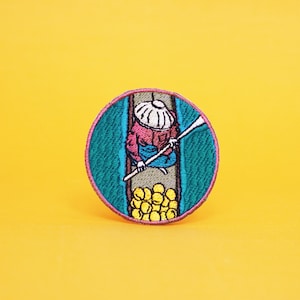 Op de afbeelding: Gestikte patch met een persoon in een boot, die peddelt met een lange roeier. De boot is gevuld met gele vruchten. De patch is rond en heeft een turquoise, roze en gele kleurstelling.