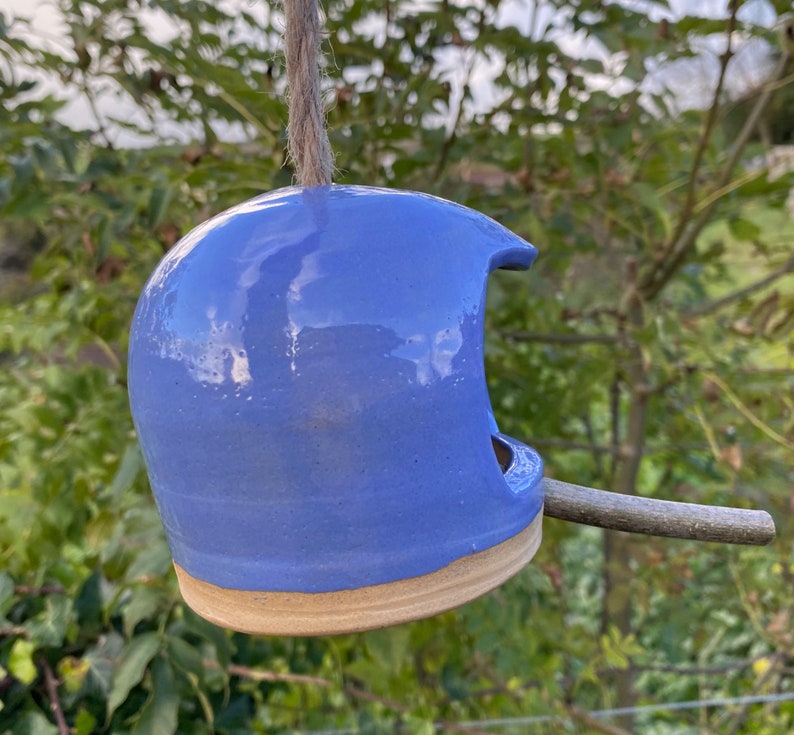 May include: A blue ceramic bird feeder with a twig perch. The feeder is hanging from a string and has a rounded shape with a hole for birds to enter.