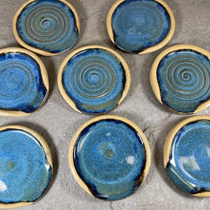 Puede incluir: Ocho posavasos de cerámica azul y beige con un diseño de espiral. Los posavasos son redondos y tienen un borde marrón claro.