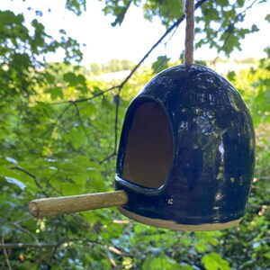 May include: A dark blue ceramic birdhouse with a small wooden perch. The birdhouse is hanging from a string and has a round opening for birds to enter.