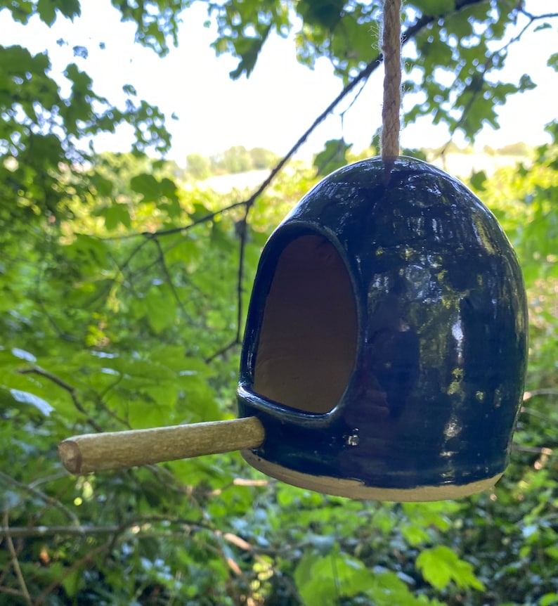 May include: A dark blue ceramic birdhouse with a small wooden perch. The birdhouse is hanging from a string and has a round opening for birds to enter.