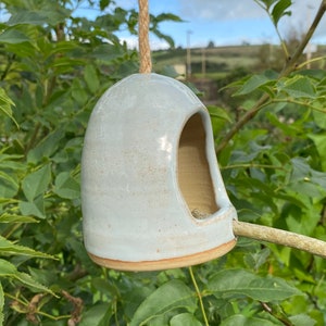 May include: A light blue ceramic bird feeder with a twig perch. The feeder is hanging from a rope and has a rounded shape with a hole for birds to enter.