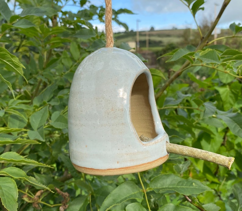 May include: A light blue ceramic bird feeder with a twig perch. The feeder is hanging from a rope and has a rounded shape with a hole for birds to enter.