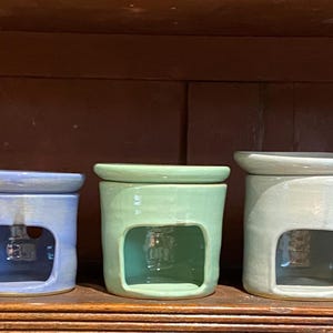 Könnte beinhalten: Fünf Keramik-Duftlampen in verschiedenen Farben: Blau, Grün, Grau und Braun. Jede Lampe hat eine quadratische Öffnung und einen Deckel. Die Lampen stehen auf einem Holzregal.