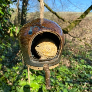 May include: A brown and blue ceramic bird feeder with a wooden perch hanging from a rope. The feeder is filled with a yellow suet cake.