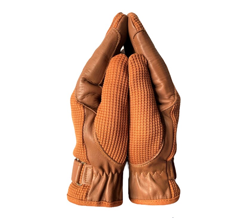May include: A pair of brown leather gloves with a textured fabric on the back of the hand. The gloves are folded together with the fingertips touching.