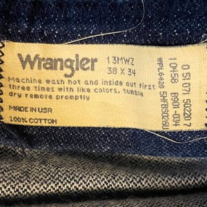 Puede incluir: Una etiqueta de cuidado de los vaqueros Wrangler de color beige con costuras negras. La etiqueta tiene el texto "Wrangler", "13MWZ", "38 X 34", "Machine wash hot and inside out first three times with like colors, tumble dry remove promptly", "MADE IN USA", "100% COTTON", "WPL6426 5HFBS0260", "10458 8901-034", "0 51 071 50220 7".