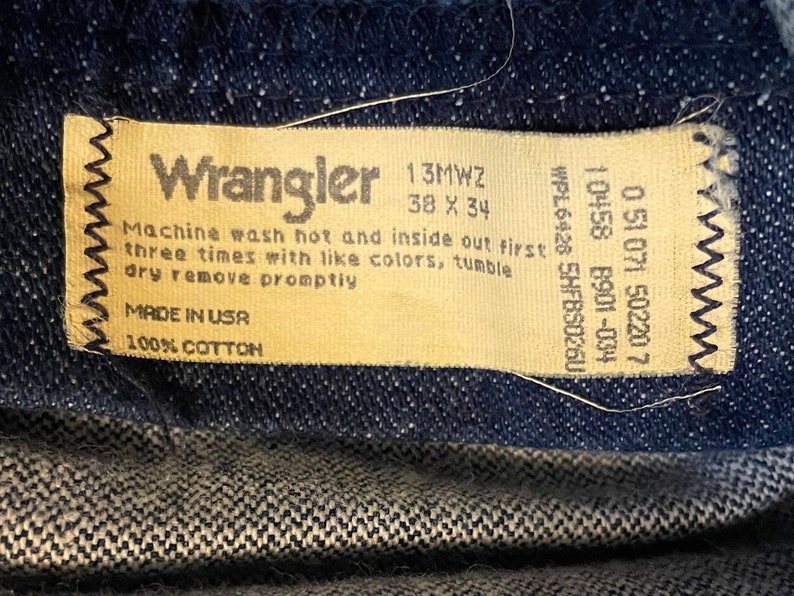 Puede incluir: Una etiqueta de cuidado de los vaqueros Wrangler de color beige con costuras negras. La etiqueta tiene el texto "Wrangler", "13MWZ", "38 X 34", "Machine wash hot and inside out first three times with like colors, tumble dry remove promptly", "MADE IN USA", "100% COTTON", "WPL6426 5HFBS0260", "10458 8901-034", "0 51 071 50220 7".