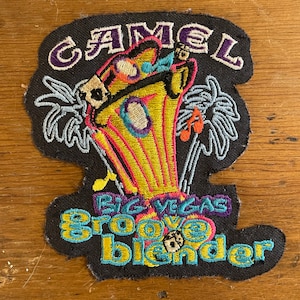 May include: A black embroidered patch with the words "Camel Big Vegas Groove Blender" in colorful lettering. The patch features a cartoon-style illustration of a person in a yellow and pink hat, holding a microphone, with palm trees in the background.