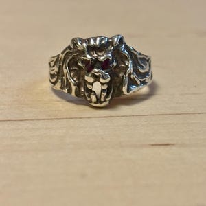 May include: A silver ring featuring a detailed lion's head design. The lion's eyes are small, red gemstones. The ring has an antique, textured finish, and is set against a light wooden surface.