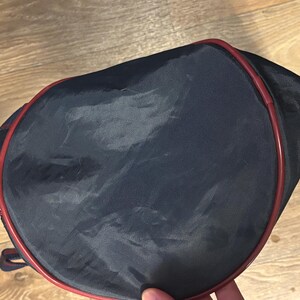May include: A navy blue tennis racket bag with a red trim. The bag is made of a shiny, possibly nylon, material. The bag is oval-shaped and has a zipper closure. The bag is designed to carry a tennis racket.