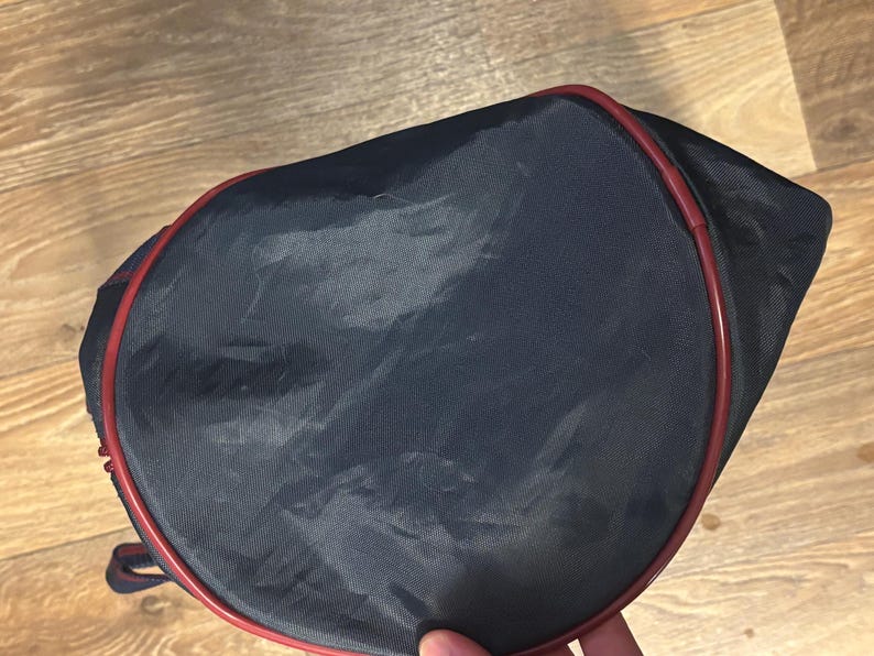 May include: A navy blue tennis racket bag with a red trim. The bag is made of a shiny, possibly nylon, material. The bag is oval-shaped and has a zipper closure. The bag is designed to carry a tennis racket.