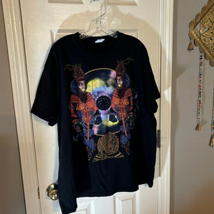 May include: Black t-shirt with a colorful graphic print of two figures wearing ornate headwear and holding a swirling orb. The figures are surrounded by a galaxy-like background.