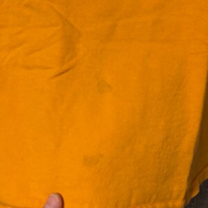 Puede incluir: Una camiseta de color amarillo mostaza s&oacute;lido hecha de una tela similar al algod&oacute;n. La prenda presenta una superficie lisa sin patrones con dos manchas tenues de forma circular visibles en la parte inferior frontal de la tela, cerca del dobladillo.