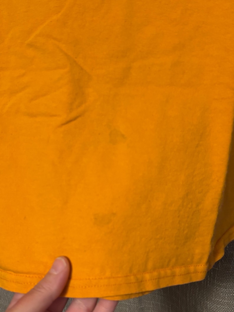 Puede incluir: Una camiseta de color amarillo mostaza s&oacute;lido hecha de una tela similar al algod&oacute;n. La prenda presenta una superficie lisa sin patrones con dos manchas tenues de forma circular visibles en la parte inferior frontal de la tela, cerca del dobladillo.