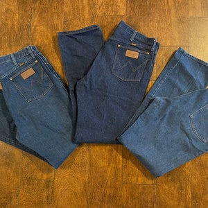 Puede incluir: Tres pares de pantalones vaqueros de mezclilla azul con un parche de cuero marr&oacute;n en el bolsillo trasero que dice "Wrangler".