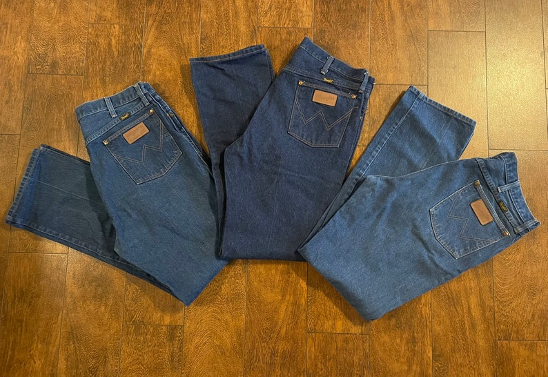Puede incluir: Tres pares de pantalones vaqueros de mezclilla azul con un parche de cuero marr&oacute;n en el bolsillo trasero que dice "Wrangler".