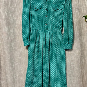 May include: A vintage teal dress with a white polka dot pattern. The dress features long sleeves, a collared neckline, and a button-down front. It has two small pockets on the chest and a gathered waist, creating a classic silhouette.