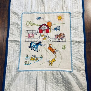 Puede incluir: Una manta de bebé acolchada blanca con un borde azul. El centro de la manta presenta una escena bordada colorida de la granja de Old MacDonald con animales y sus sonidos.