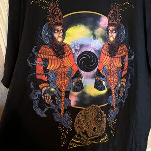 May include: Black t-shirt with a graphic print of two figures wearing ornate headwear and robes, holding orbs in their hands. The figures are standing in front of a swirling, colorful background with a black circle in the center. A golden bear is depicted at the bottom of the design.