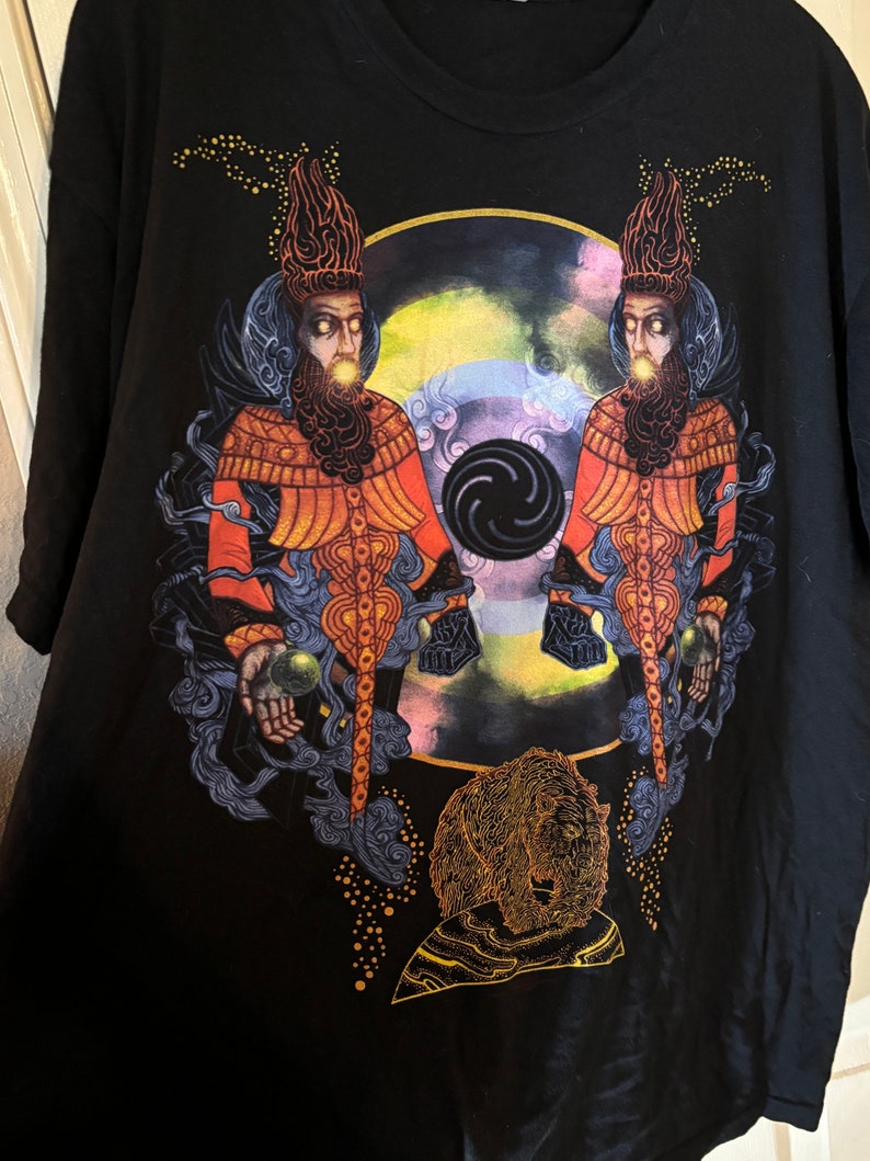 May include: Black t-shirt with a graphic print of two figures wearing ornate headwear and robes, holding orbs in their hands. The figures are standing in front of a swirling, colorful background with a black circle in the center. A golden bear is depicted at the bottom of the design.