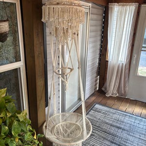 May include: A cream-colored macrame hanging chair featuring an intricate tiered canopy with knotted fringe and wooden beads. The structure includes a circular seat base with a woven cord pattern, suspended by thick braided ropes from a central top point.