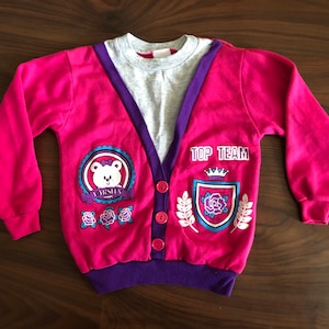 Puede incluir: Sudadera rosa de manga larga con ribete morado y una camiseta interior gris. La sudadera tiene un gráfico de oso con el texto "Varsity" y un escudo con el texto "Top Team".