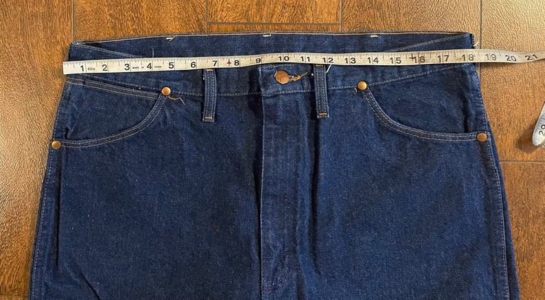 Puede incluir: Un par de pantalones vaqueros de mezclilla azul oscuro con un ojal para cintur&oacute;n de cuero marr&oacute;n y un cierre de bot&oacute;n met&aacute;lico. Los pantalones vaqueros se miden con una cinta m&eacute;trica, mostrando una talla de cintura de 46 cm.