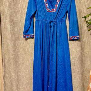 May include: A long, vibrant blue robe with long sleeves. The robe features a V-neckline and cuffs adorned with a floral pattern in red, white, and blue. A matching tie cinches the waist. The robe is hanging on a hanger.