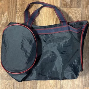 May include: A black tote bag with a matching oval-shaped side pocket. The bag has red trim and navy blue and red striped handles. The bag is made of a shiny, possibly nylon, material.