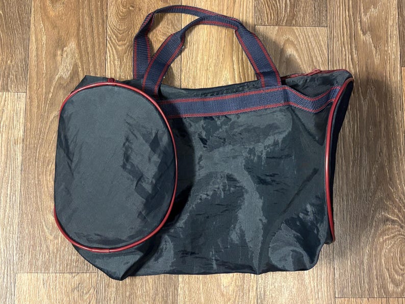 May include: A black tote bag with a matching oval-shaped side pocket. The bag has red trim and navy blue and red striped handles. The bag is made of a shiny, possibly nylon, material.