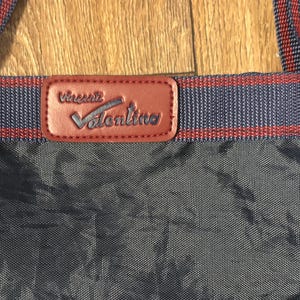 May include: Close-up of a navy blue tote bag with red and navy blue striped straps. A brown leather patch with the name "Vincent Valentino" is centered on the bag.