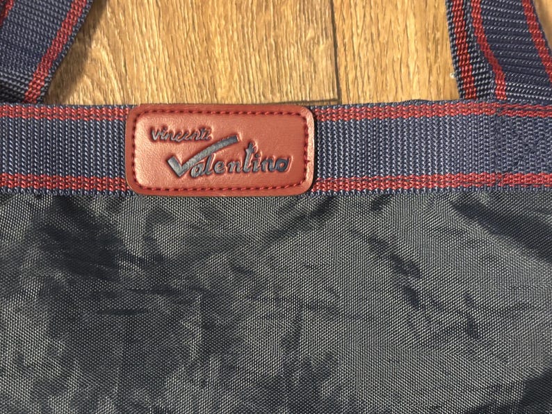 May include: Close-up of a navy blue tote bag with red and navy blue striped straps. A brown leather patch with the name "Vincent Valentino" is centered on the bag.