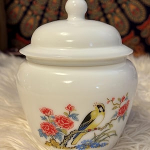 May include: A white ceramic jar with a lid, featuring a painted design of a yellow bird perched on a branch with red flowers and blue accents. The jar has a glossy finish and a decorative finial on the lid.