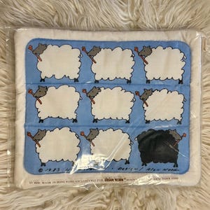 May include: A packaged rectangular item with a blue background and white sheep illustrations. Each sheep holds a red-tipped stick. One sheep is black. Text reads "© 1983 Lillian Vernon Design / Alan Wood".