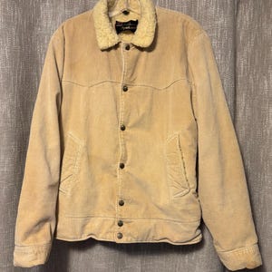 May include: A tan corduroy jacket with a cream shearling collar. The jacket features a button-up front, two front pockets, and buttoned cuffs. The jacket is suspended from a hanger.