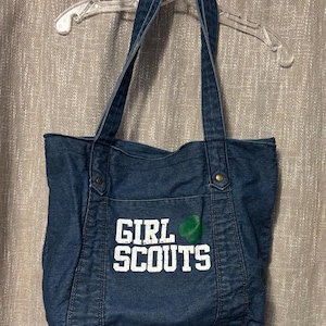 May include: A denim tote bag with long straps, featuring the words "GIRL SCOUTS" in white block letters on the front. A green emblem is next to the text. The bag is hanging on a clear plastic hanger.