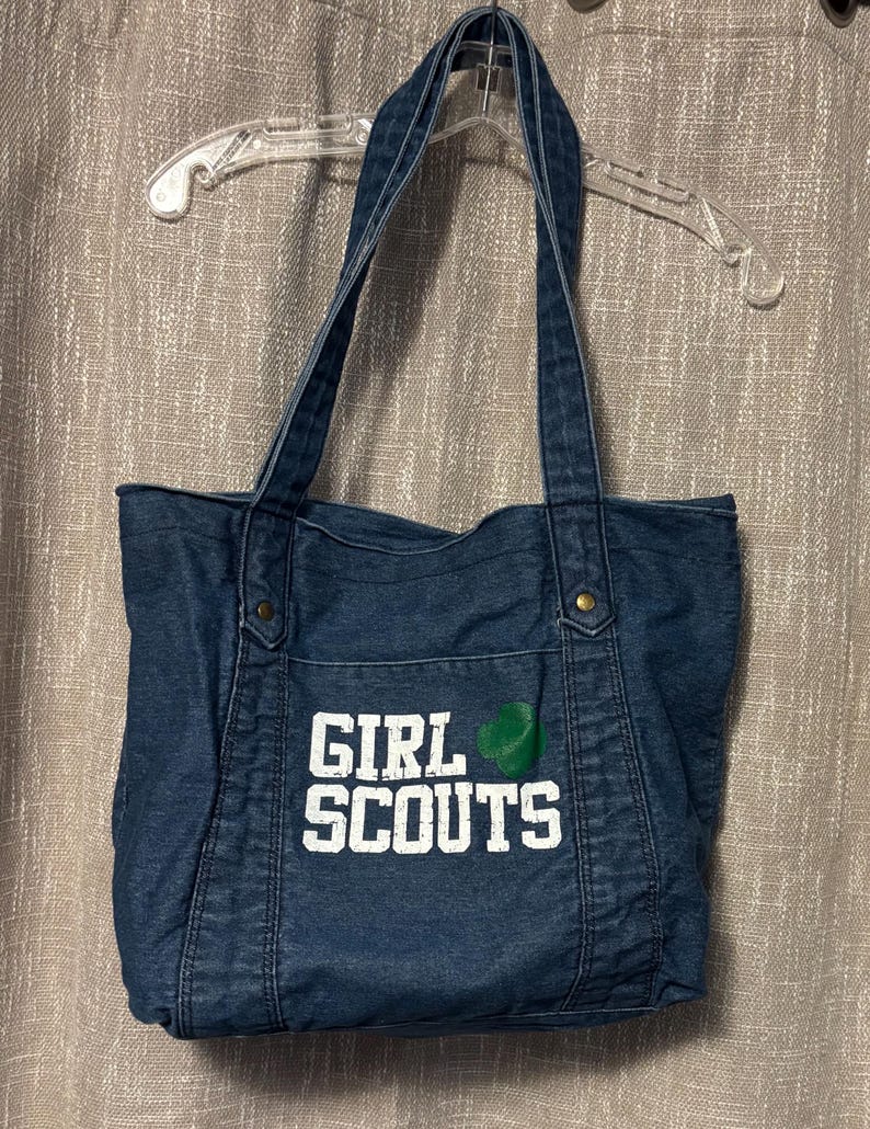 May include: A denim tote bag with long straps, featuring the words "GIRL SCOUTS" in white block letters on the front. A green emblem is next to the text. The bag is hanging on a clear plastic hanger.
