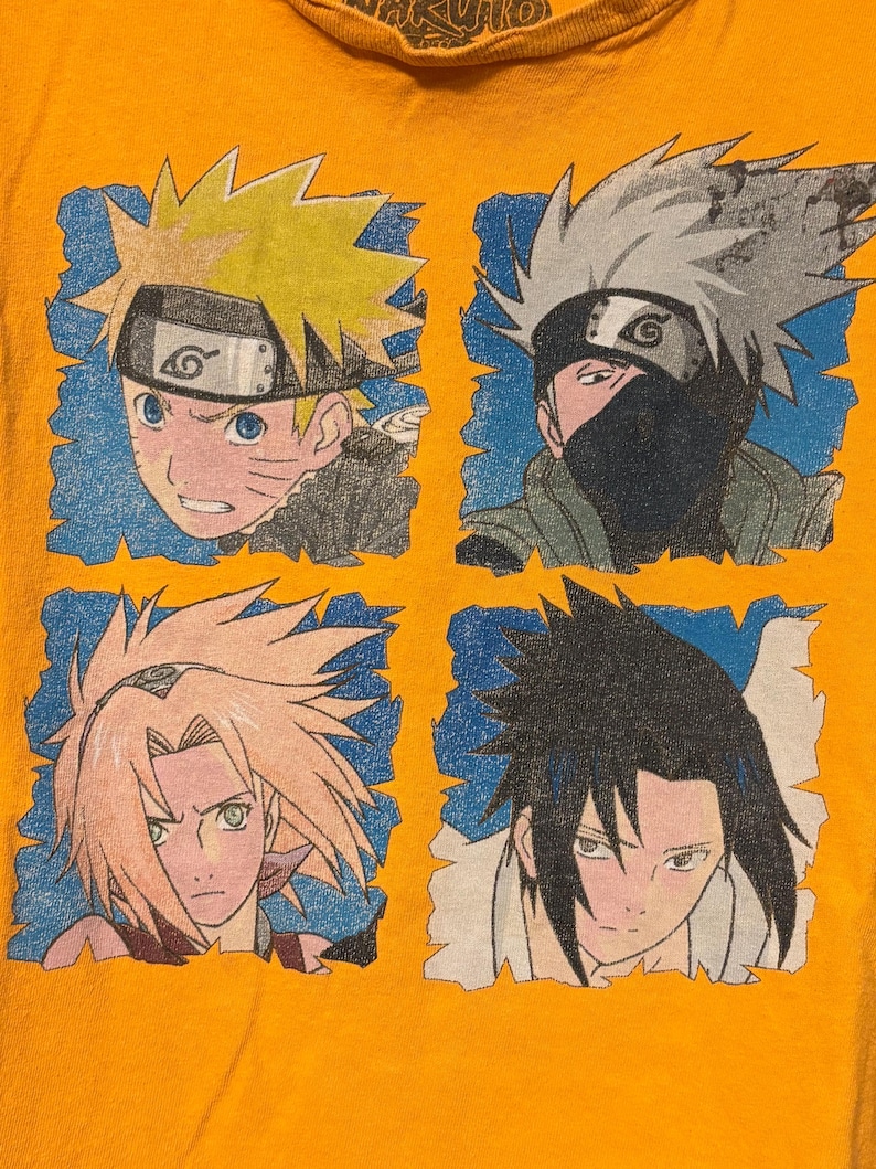 Puede incluir: Una camiseta naranja con un estampado gr&aacute;fico de cuatro personajes de anime de Naruto. El dise&ntilde;o muestra a Naruto, Kakashi, Sakura y Sasuke, cada uno enmarcado en un cuadrado azul. El texto Naruto es visible en la parte superior del cuello.