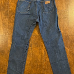 Puede incluir: Un par de pantalones vaqueros de mezclilla azul oscuro con un parche de cuero marr&oacute;n en el bolsillo trasero que dice "Wrangler".