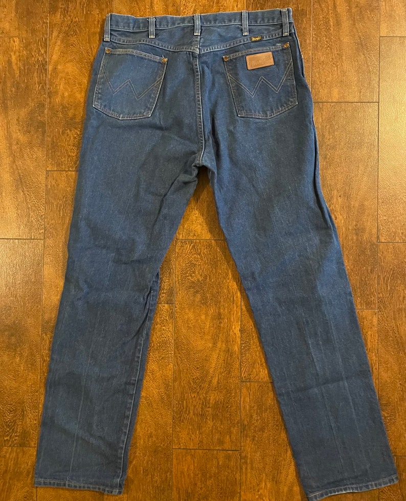 Puede incluir: Un par de pantalones vaqueros de mezclilla azul oscuro con un parche de cuero marr&oacute;n en el bolsillo trasero que dice "Wrangler".