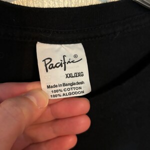 May include: Black t-shirt with a white tag that says "Pacific" and "XXL/2XG" in black text. The tag also says "Made in Bangladesh 100% Cotton 100% Algodon".