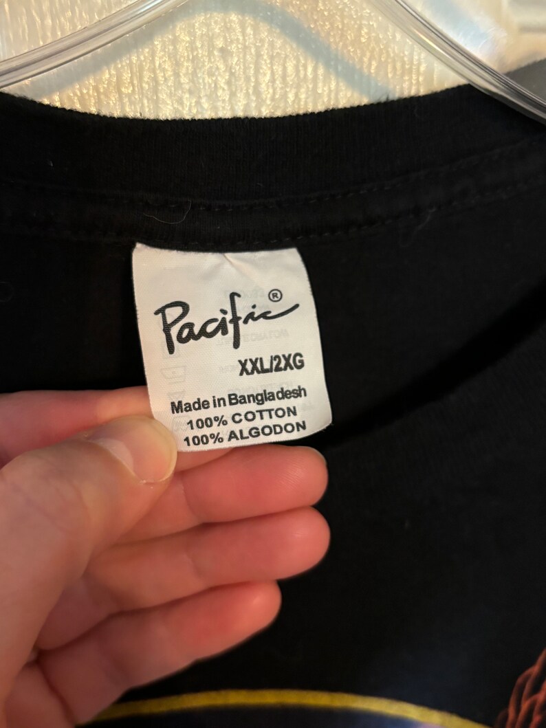 May include: Black t-shirt with a white tag that says "Pacific" and "XXL/2XG" in black text. The tag also says "Made in Bangladesh 100% Cotton 100% Algodon".