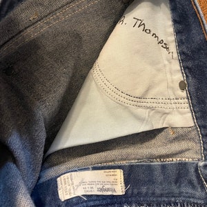 Puede incluir: Primer plano del interior de un par de pantalones vaqueros azules. Los vaqueros tienen una etiqueta de Wrangler y un nombre escrito a mano, "R. Thompson", dentro del bolsillo trasero.