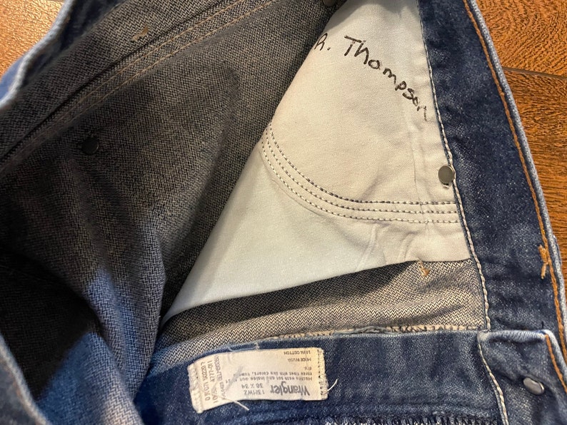 Puede incluir: Primer plano del interior de un par de pantalones vaqueros azules. Los vaqueros tienen una etiqueta de Wrangler y un nombre escrito a mano, "R. Thompson", dentro del bolsillo trasero.