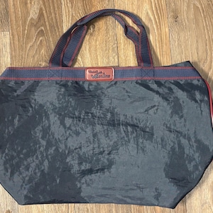 May include: A navy blue tote bag with red trim and handles. The bag has a rectangular shape with a slightly rounded bottom. The handles are navy blue with red stripes. The top of the bag has a red stripe with a navy blue stripe above it.