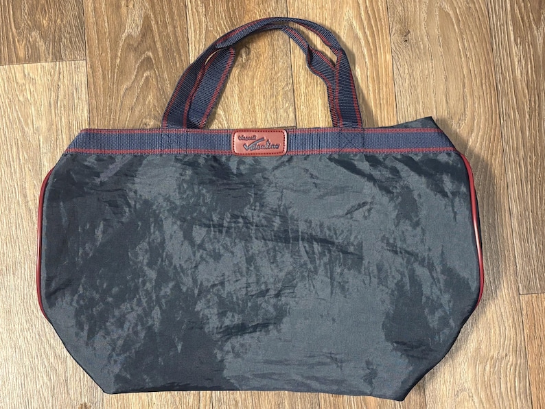 May include: A navy blue tote bag with red trim and handles. The bag has a rectangular shape with a slightly rounded bottom. The handles are navy blue with red stripes. The top of the bag has a red stripe with a navy blue stripe above it.